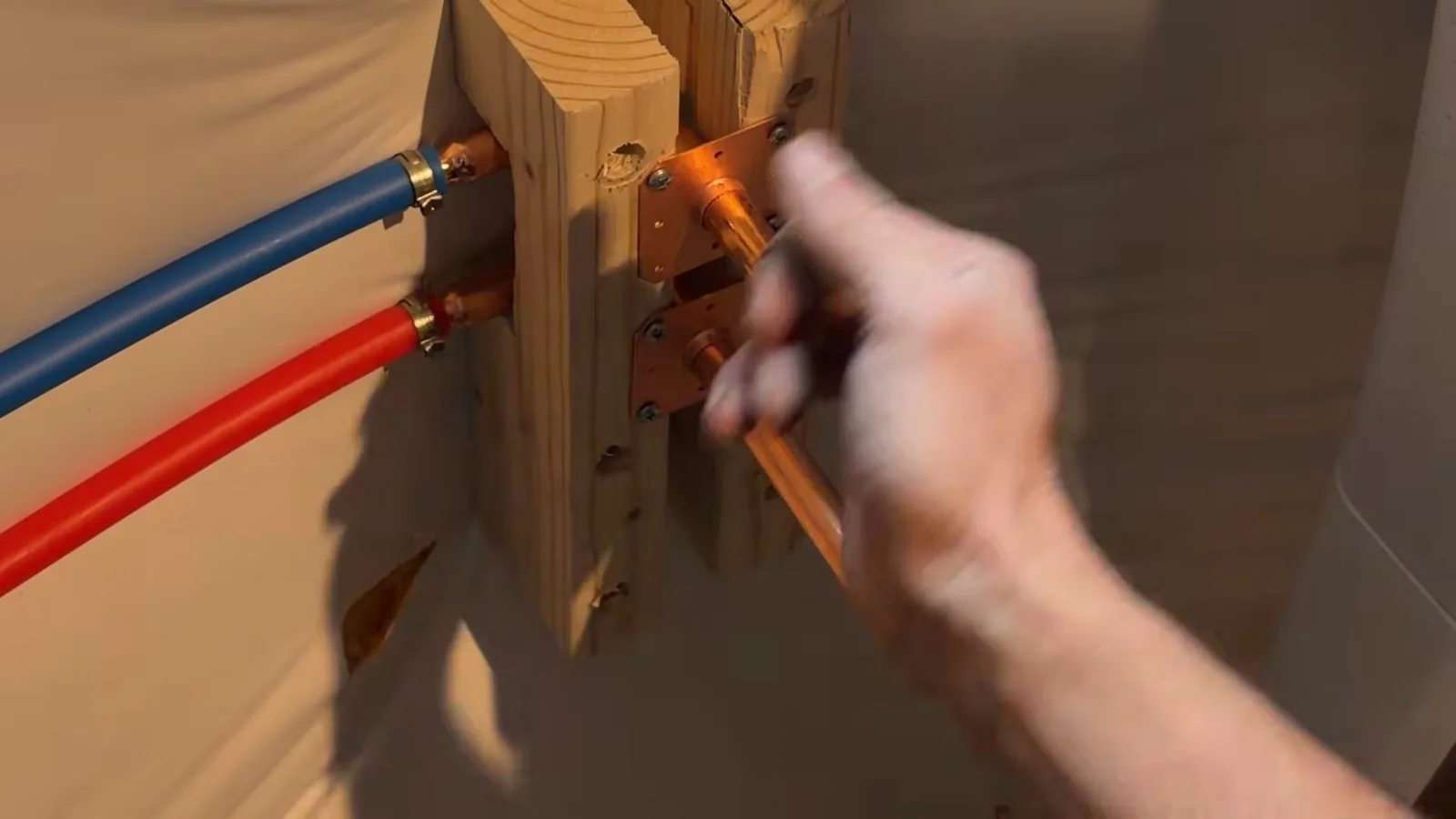 A hand fitting a copper elbow beside blue and red PEX lines mounted on a wooden block