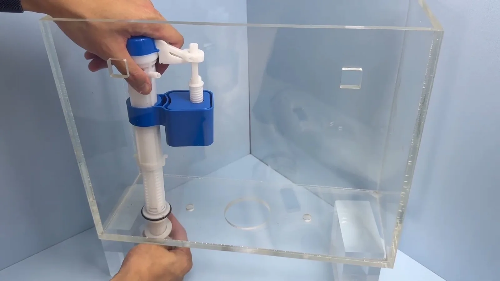 Two hands setting a blue fill valve into a clear acrylic toilet tank