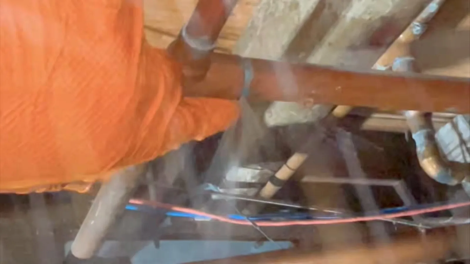A gloved hand on a copper pipe in a crawlspace with water spraying from the break