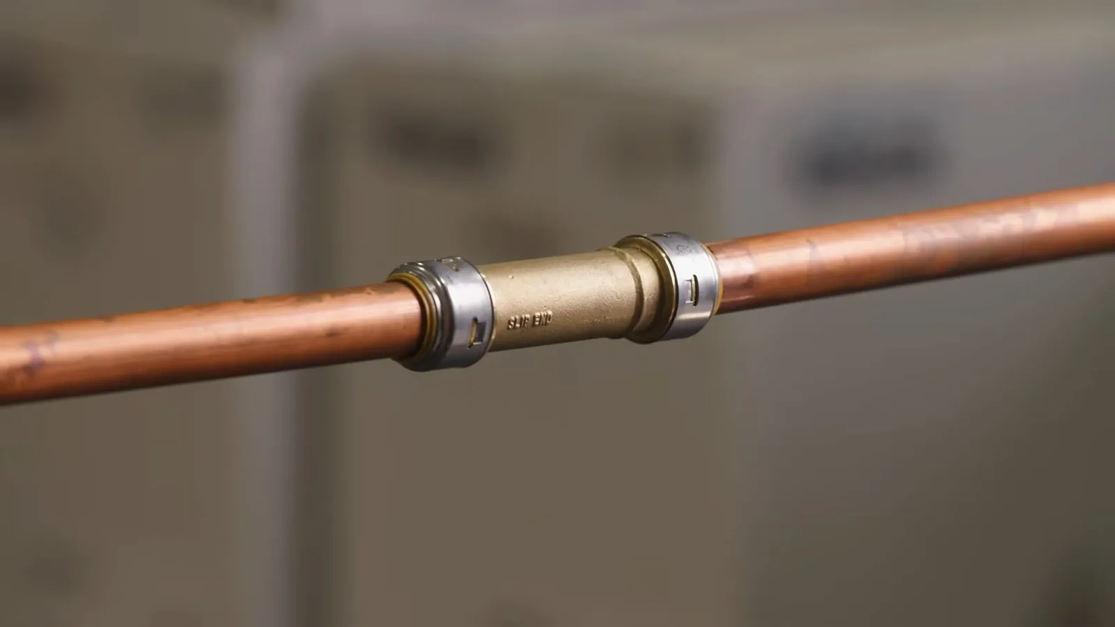 A copper pipe joined by a brass coupling with a blurred room behind it