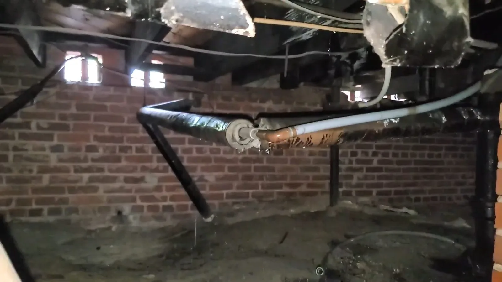 Black and gray pipes running beneath a floor in a brick-walled crawlspace