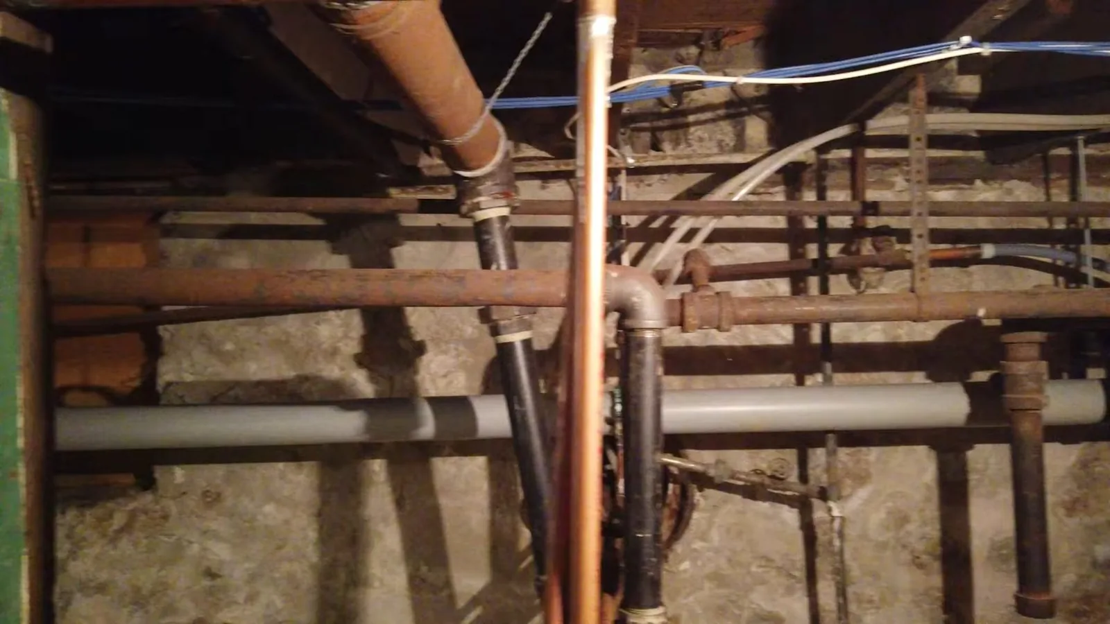 Rusted and copper pipes running through an unfinished basement ceiling between wood joists