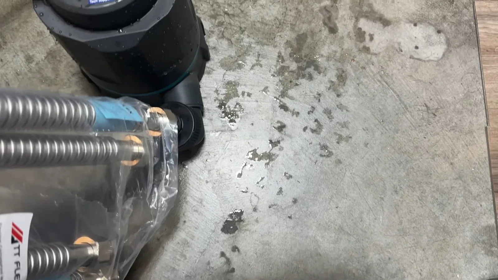 Flexible stainless hoses lying on a concrete floor beside the base of a dark water heater