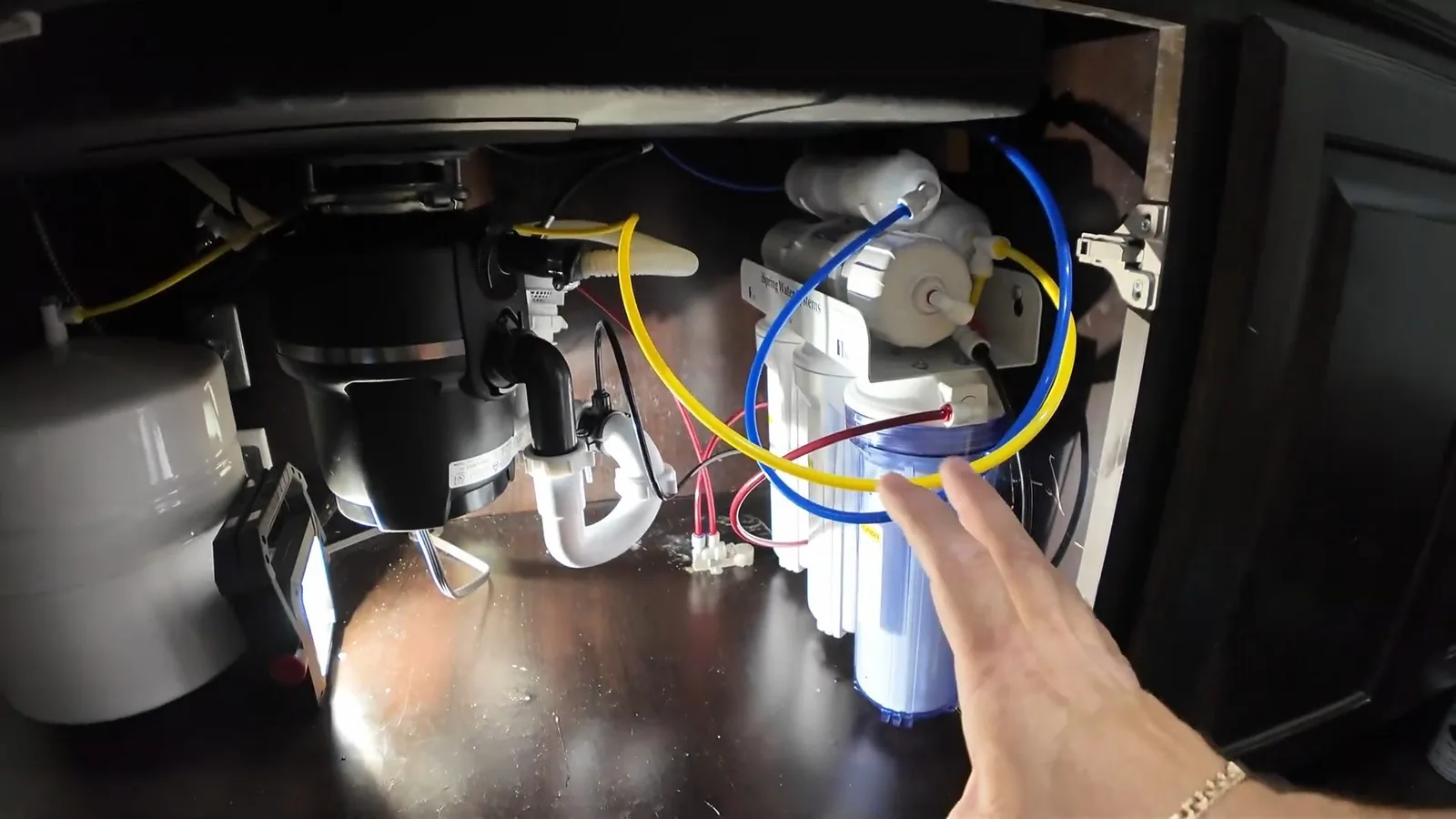 Inside an under-sink cabinet, a garbage disposal and water filter with colored tubing while a hand reaches in