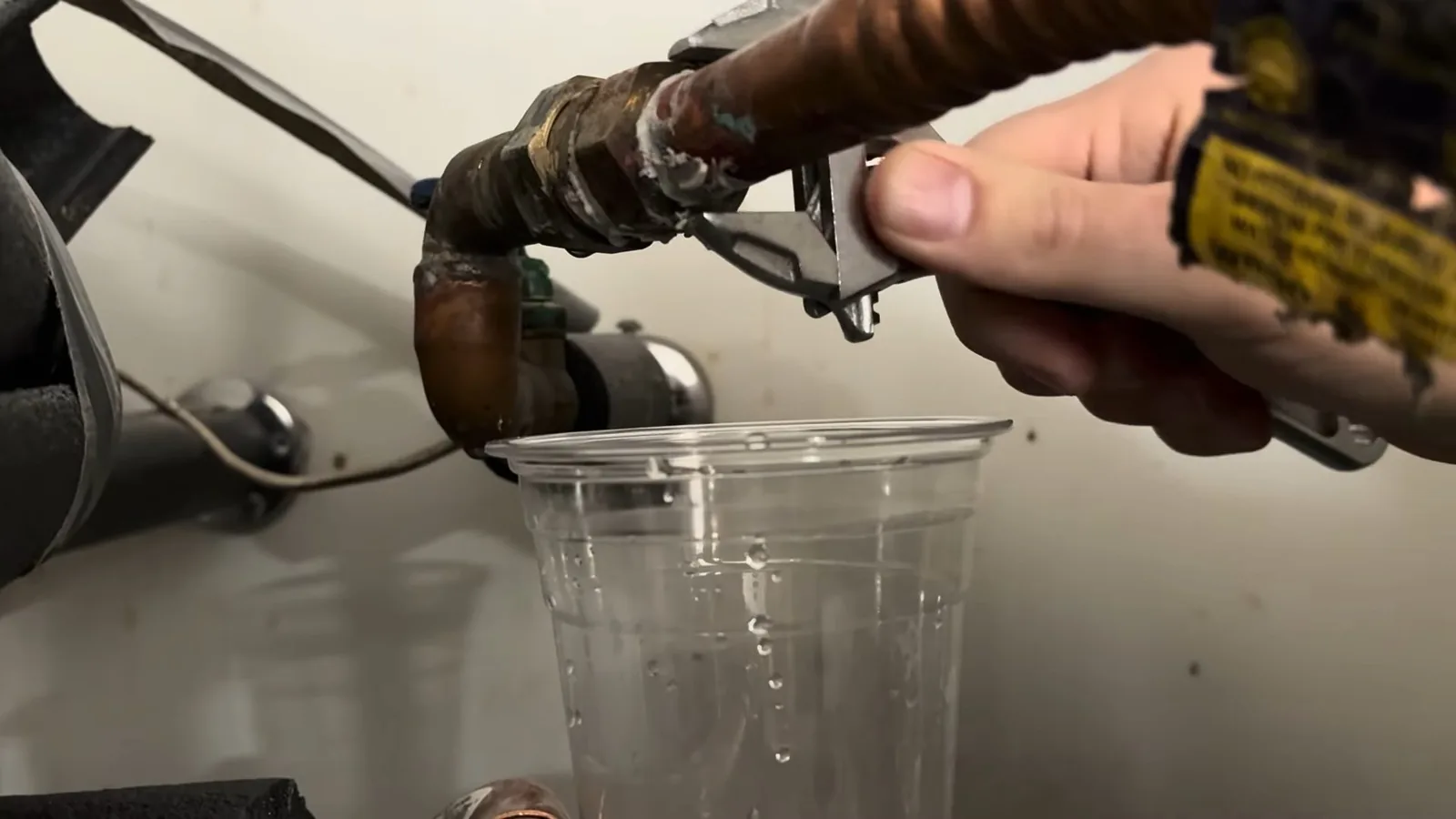A hand holding a wrench on a brass fitting above a clear plastic cup catching drips