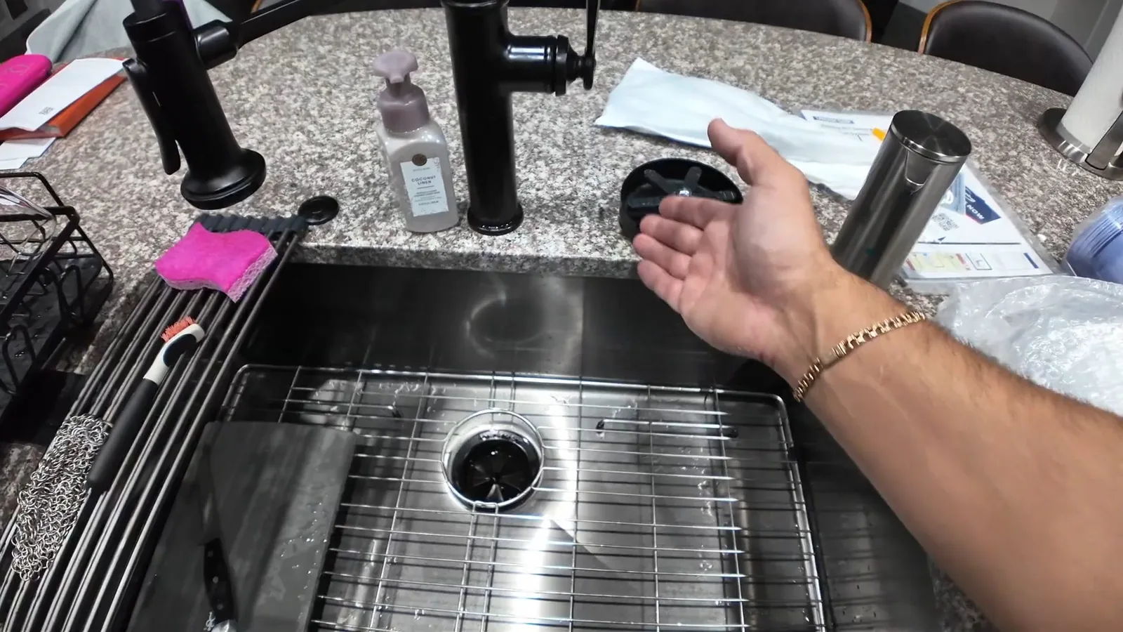 A hand held over a stainless steel kitchen sink with a drain opening, black faucets and a soap dispenser