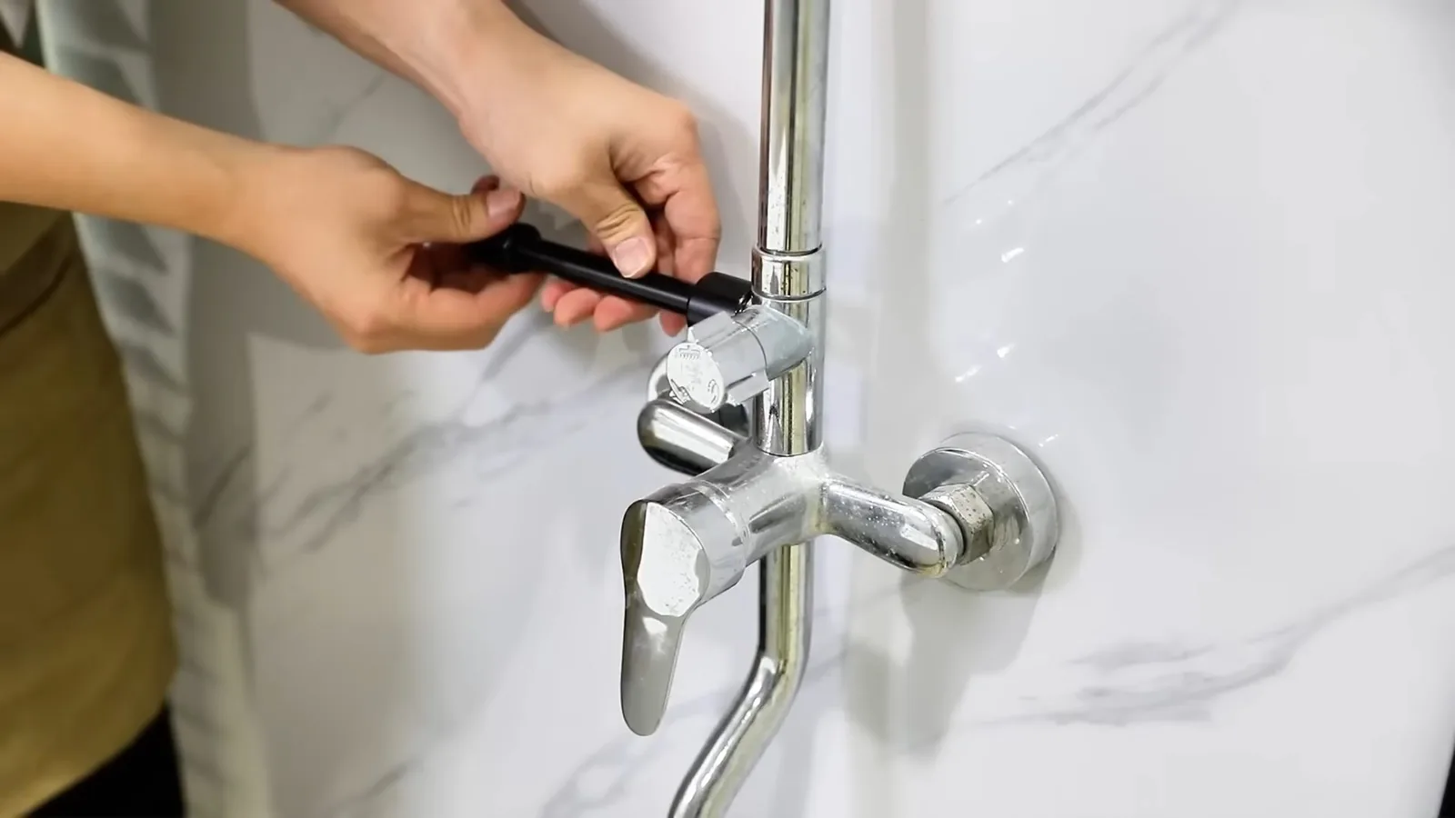 Hands adjusting the handle of a chrome wall-mounted faucet against a white marble-look wall