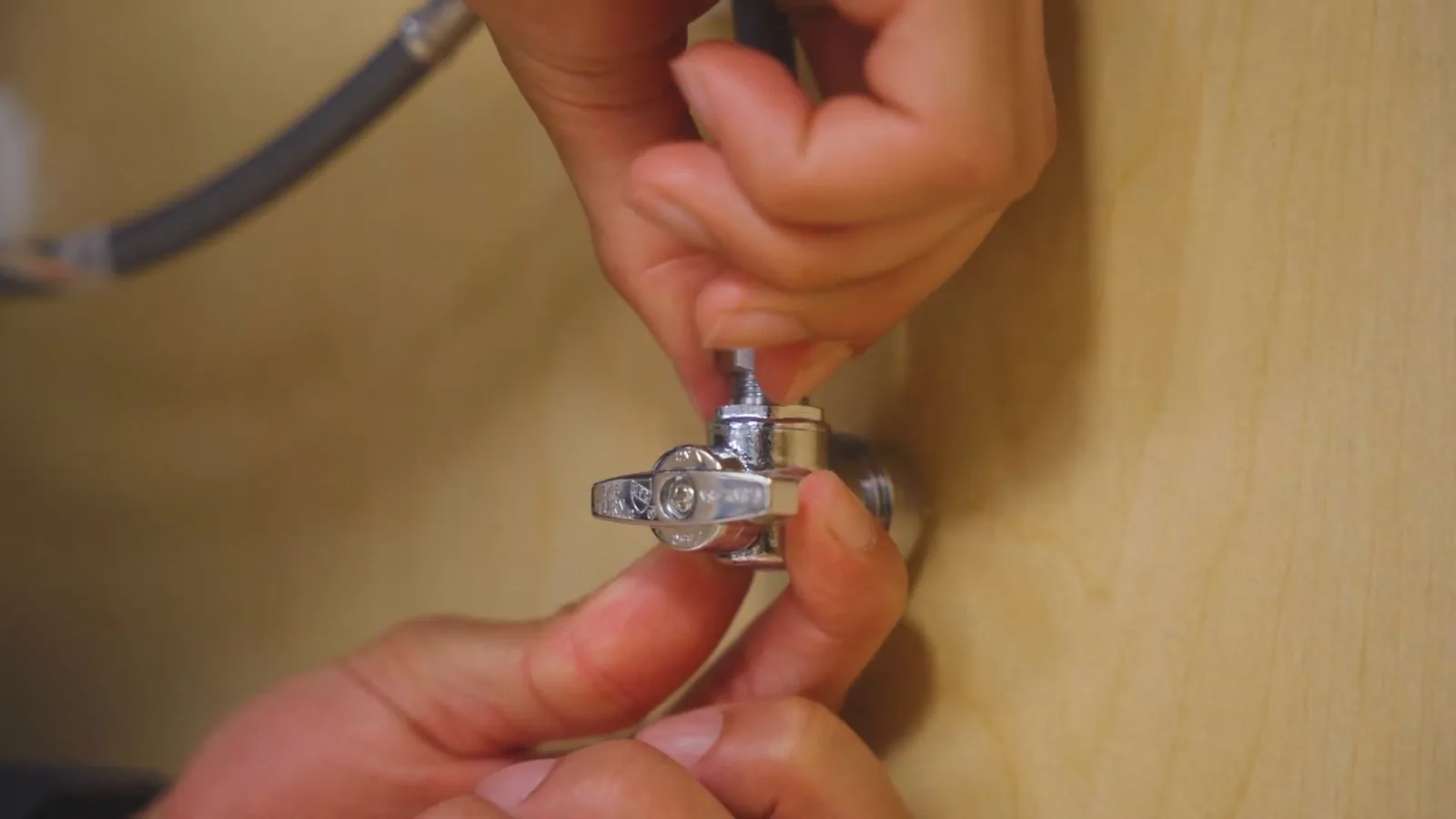 Two hands turning a small chrome shutoff valve against a light wood panel