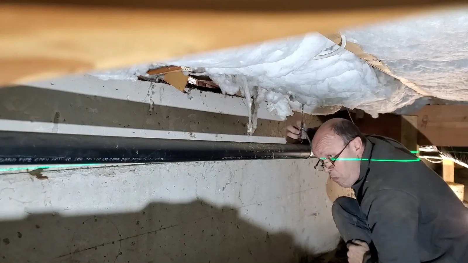 A man crouching in a low crawlspace beneath a floor, looking at a pipe run