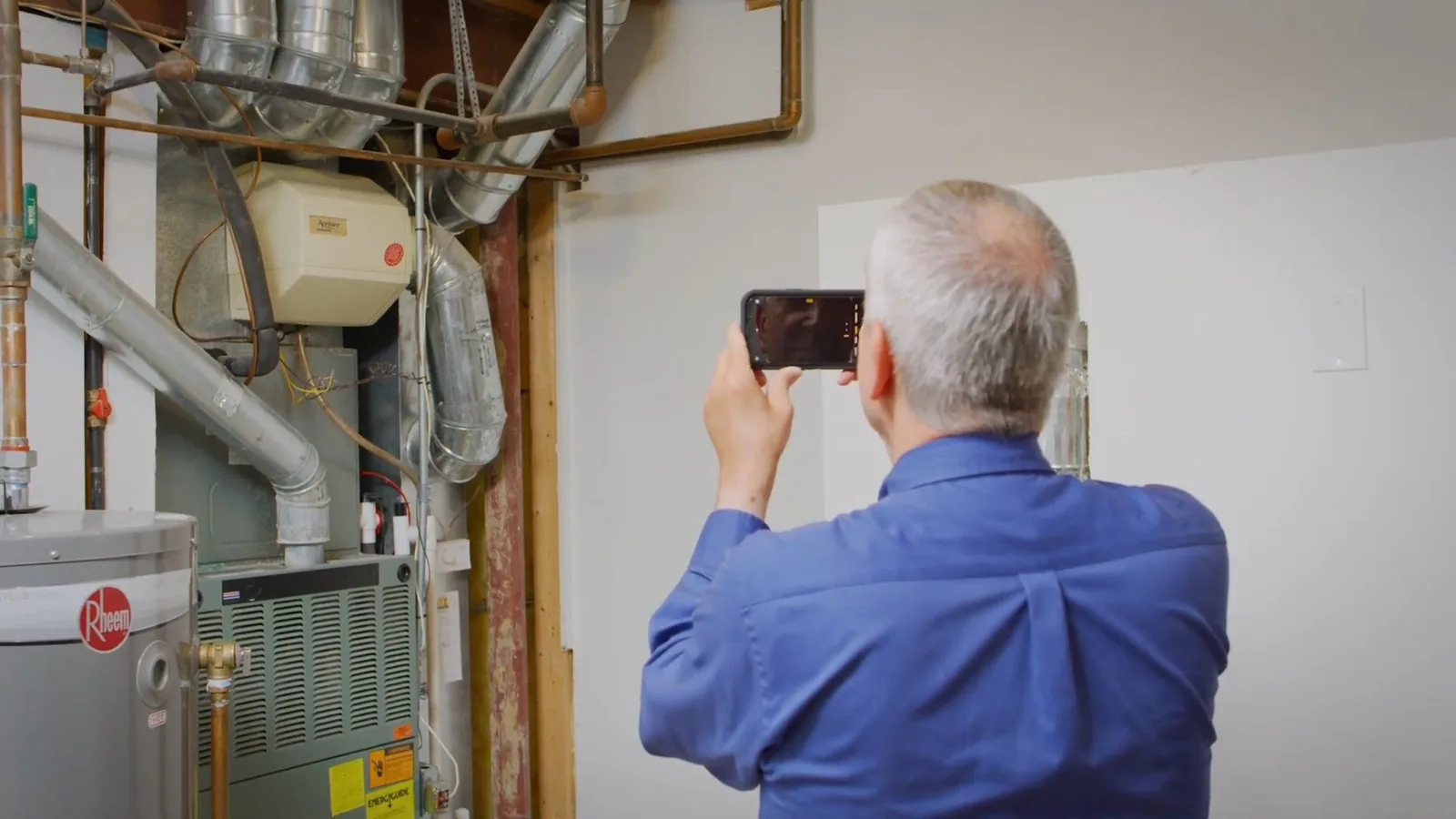 A man in a blue shirt photographing a water heater with a phone in a utility closet beside a furnace