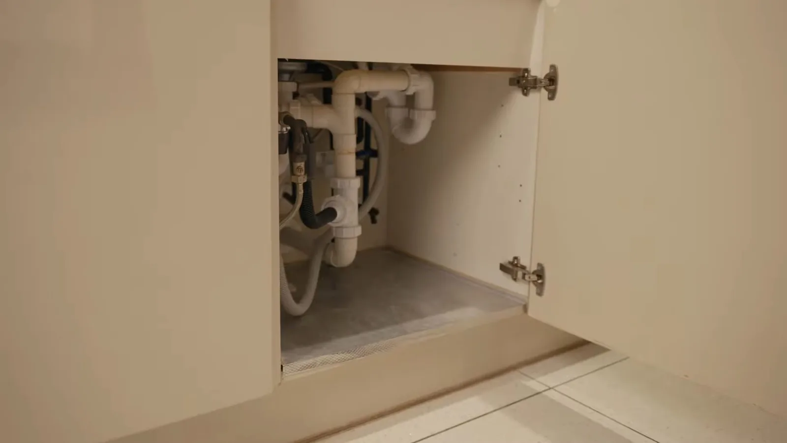 An open cabinet door showing white drain pipes and supply lines under a sink