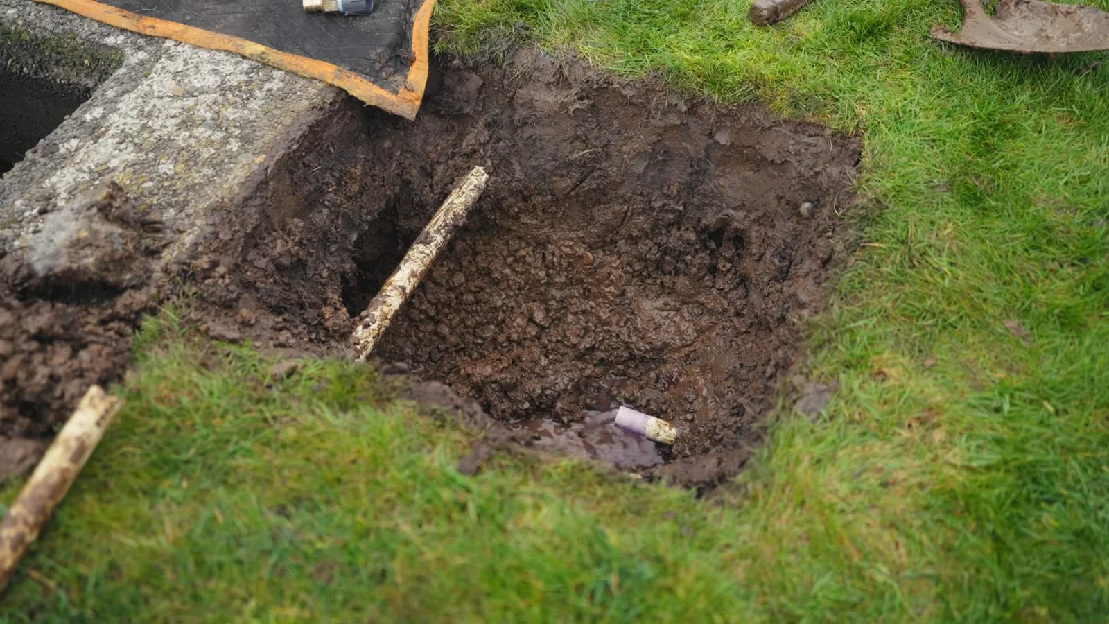 A square hole dug in a lawn beside a stone edge, with a wooden stake and a short pipe in the soil