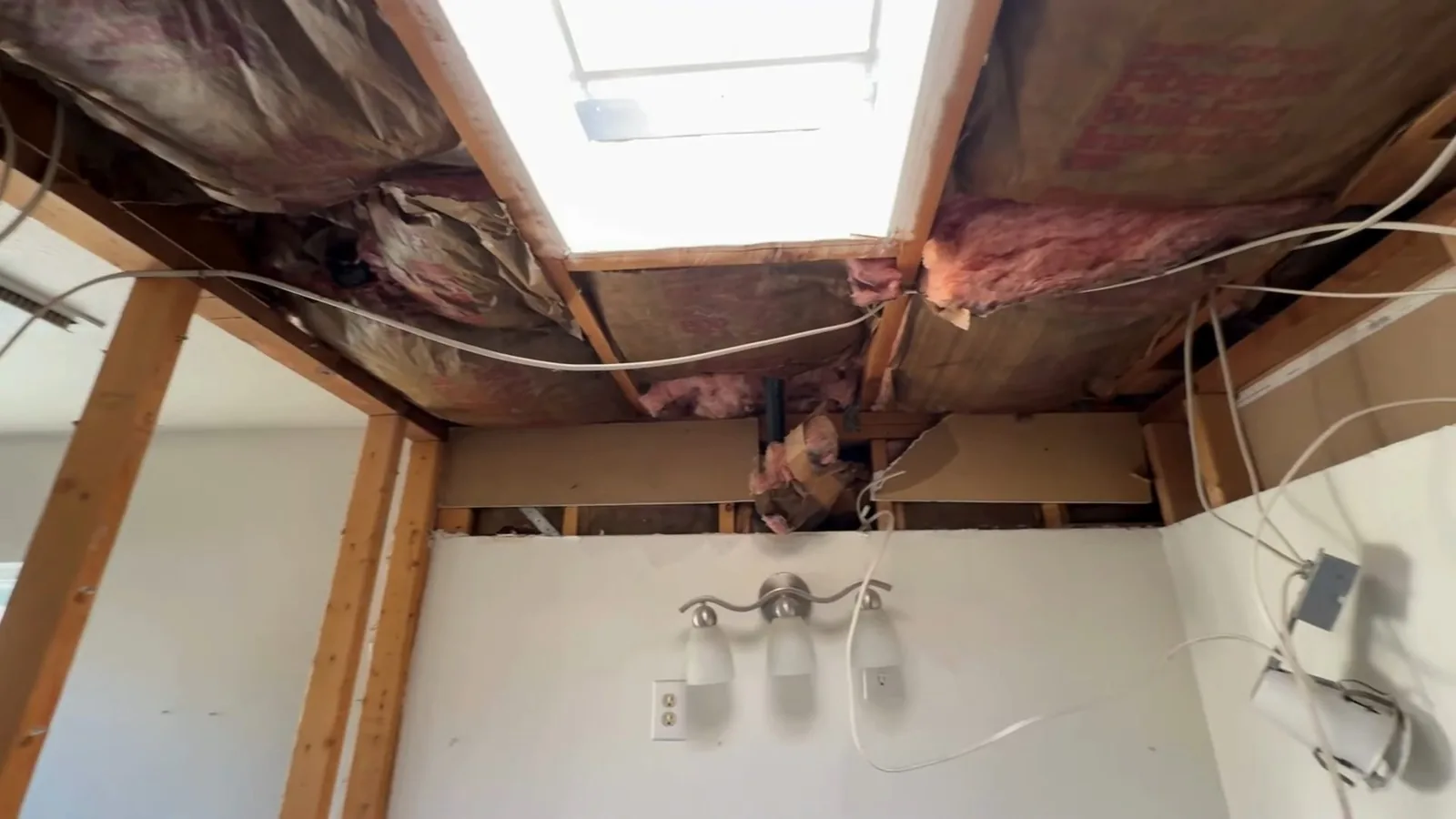 An opened bathroom ceiling with exposed joists, pink insulation and wiring above a vanity light