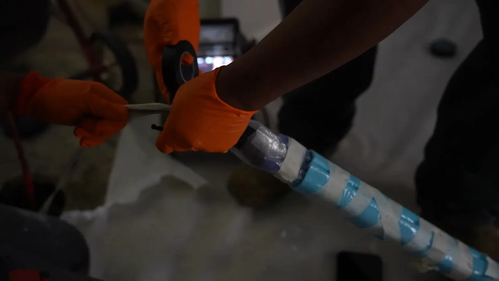 Orange-gloved hands wrapping a long tube with blue and white tape in dim light