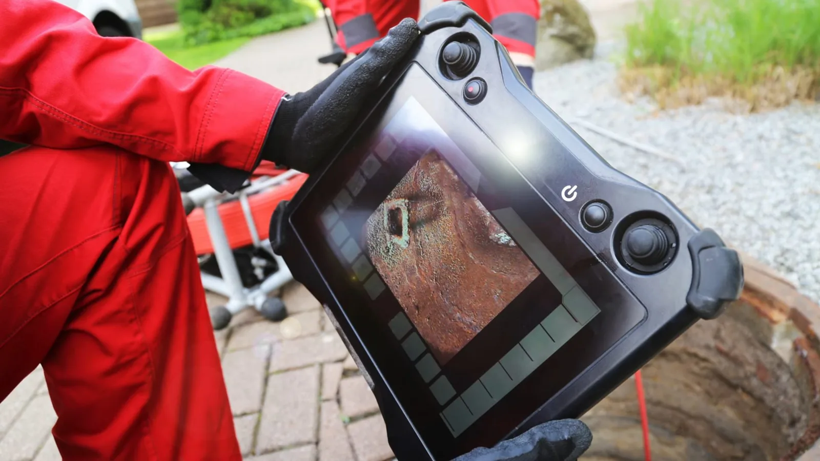 A person in red coveralls holding a handheld sewer camera monitor on a brick-paved patio