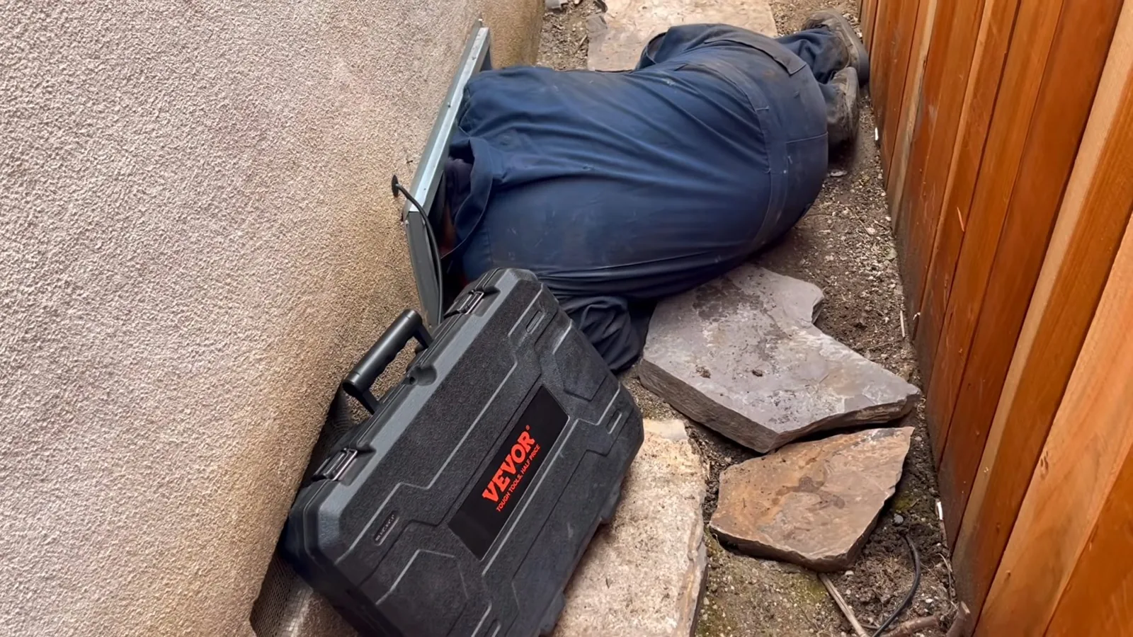 A plumber lying on the ground beside a stucco wall and fence, working at a low opening next to a tool case