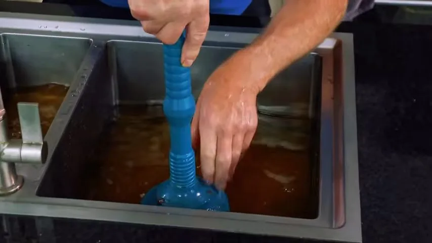 Two hands working a blue plunger in a stainless steel sink full of dirty water
