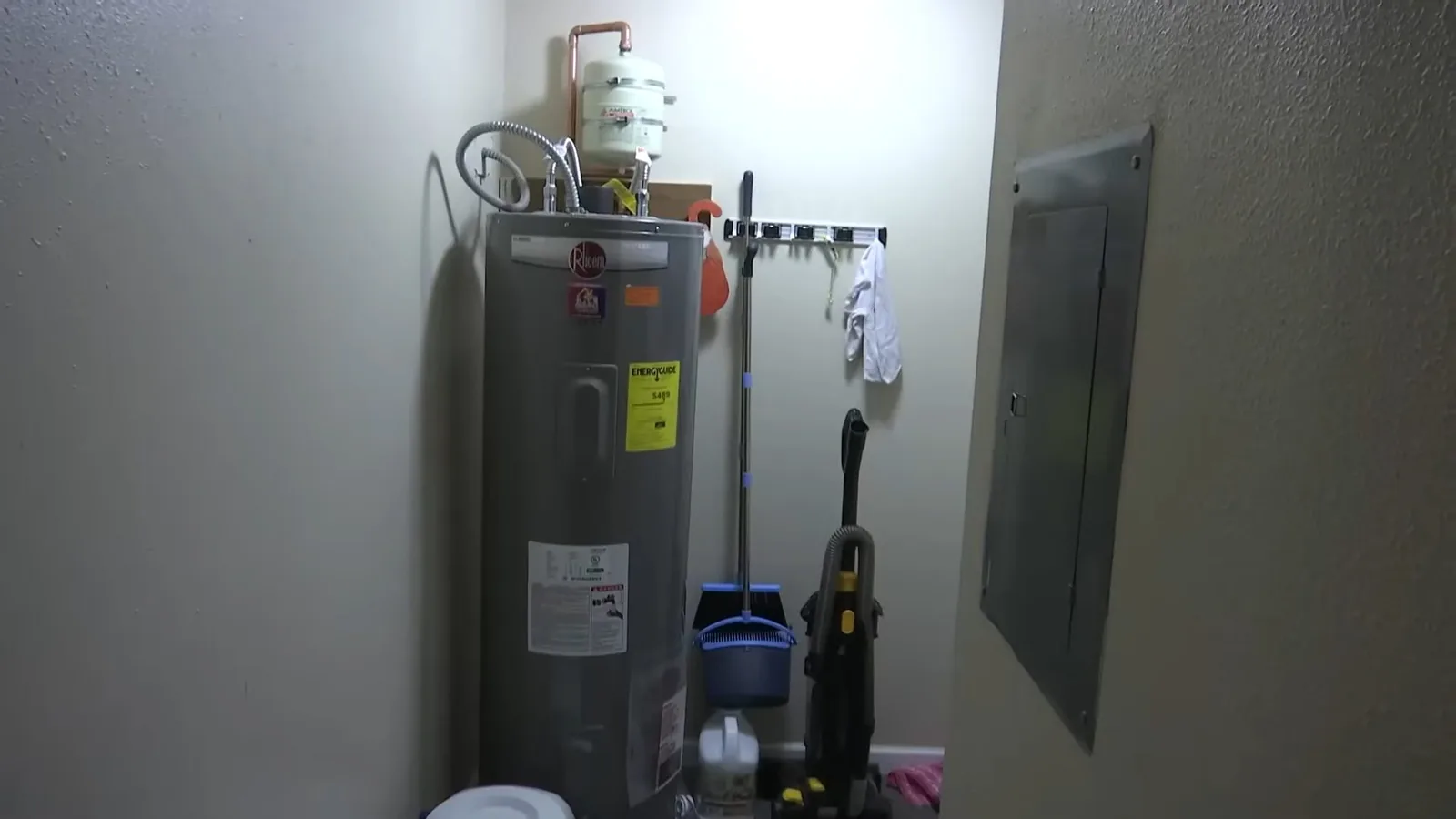A tall tank water heater against a wall in a utility room beside a mop, a dustpan and an electrical panel