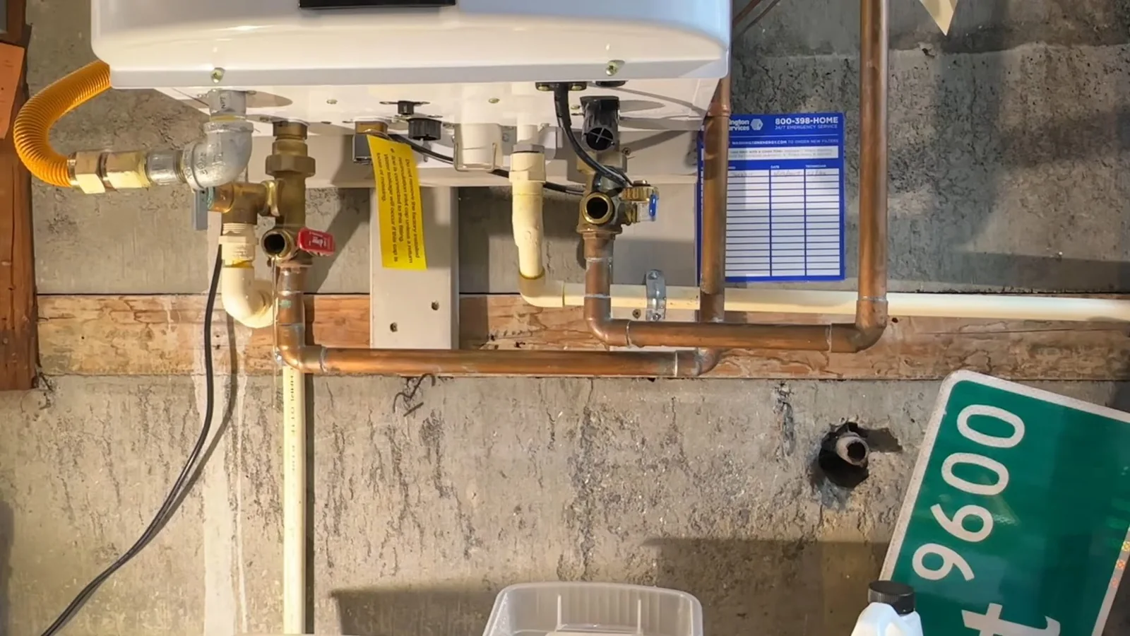 A white tankless water heater above brass valves, a yellow gas line and copper pipes against a concrete wall