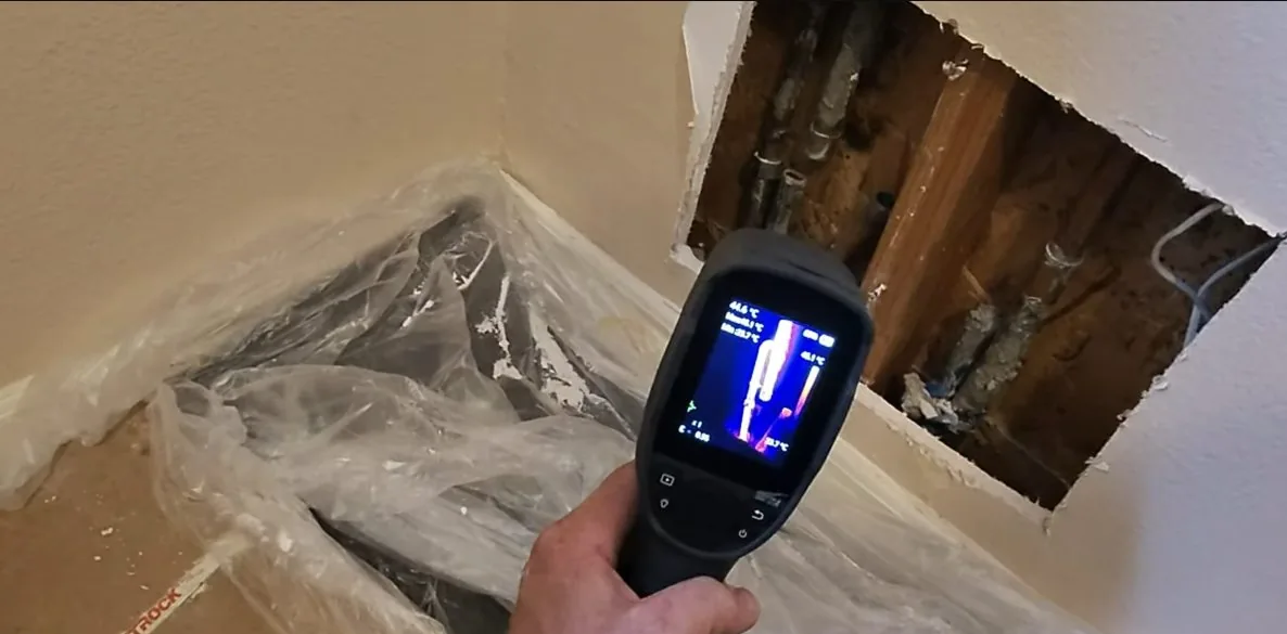 A handheld thermal leak detector held toward an opened wall corner with plastic sheeting on the floor