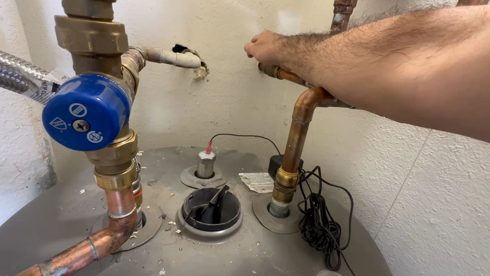 A forearm reaching over the top of a water heater with brass fittings, a blue valve and a drain pipe