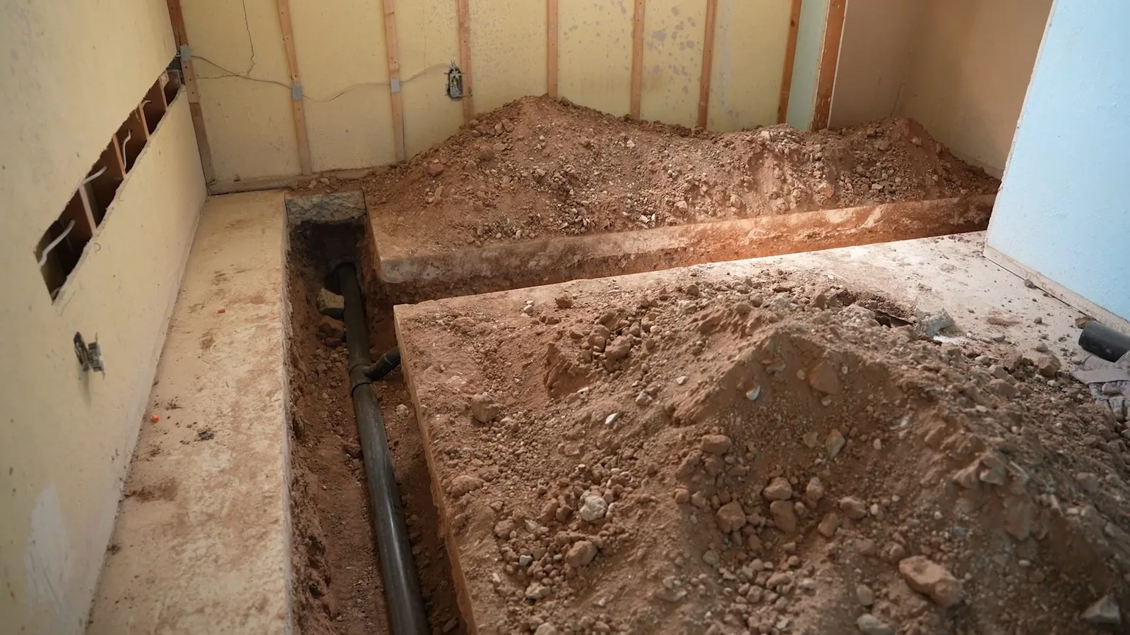 A trench cut in a basement concrete floor with piles of soil beside it and framed walls around