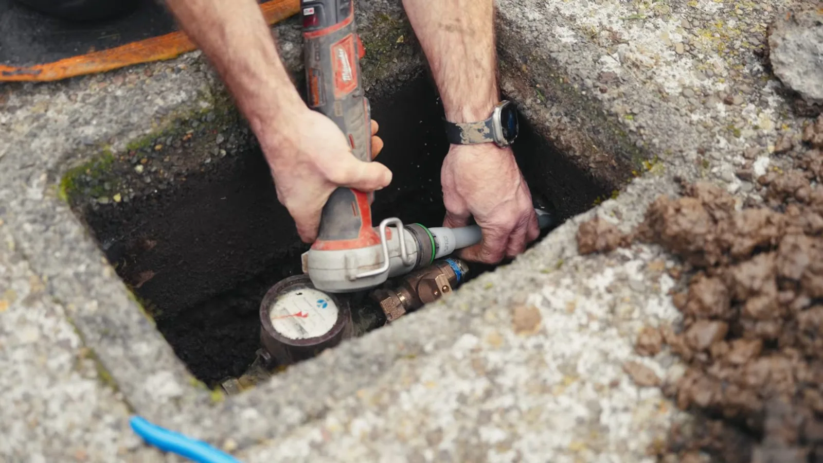Hands using a power tool on a pipe beside a water meter inside a box set in the ground