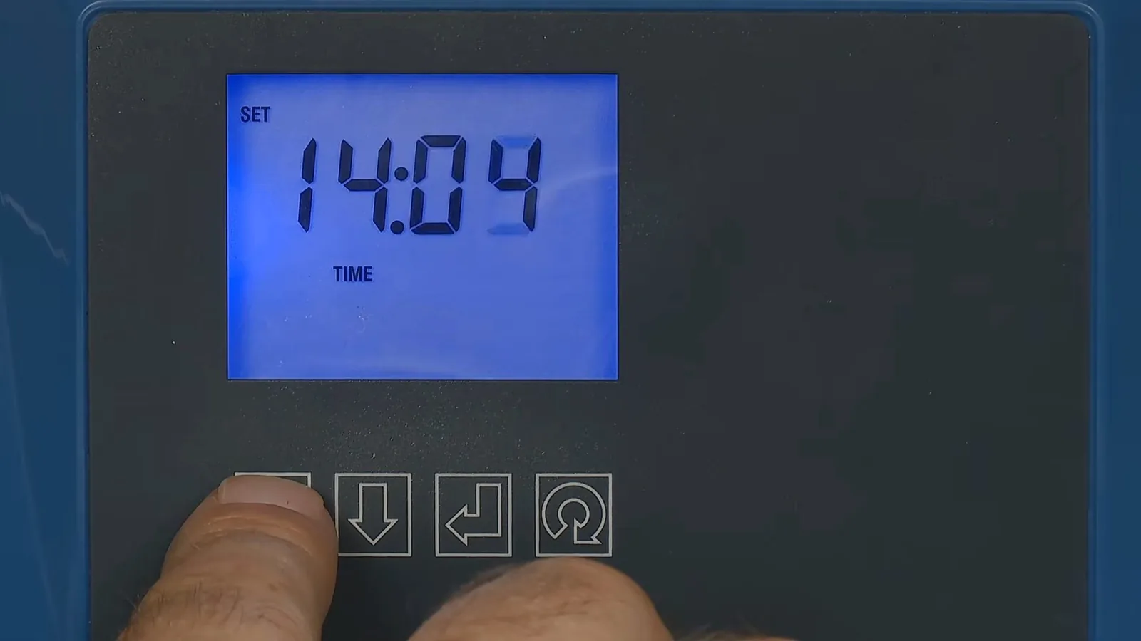 A blue backlit time display and four control buttons on a water softener head, with a thumb pressing one