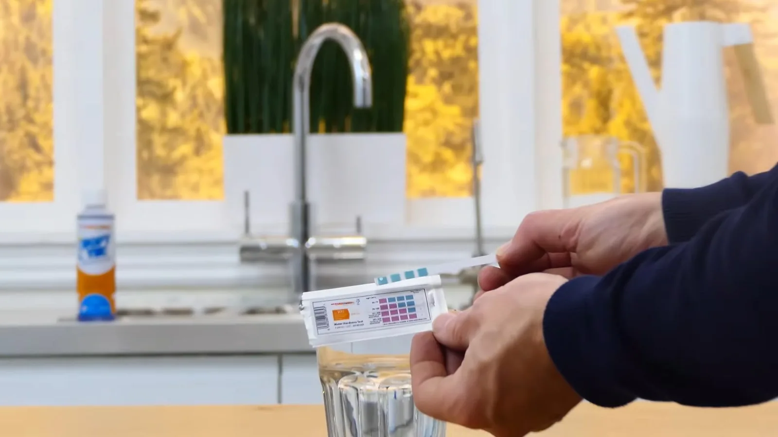 A hand dipping a water test strip into a glass of tap water beside a kitchen faucet