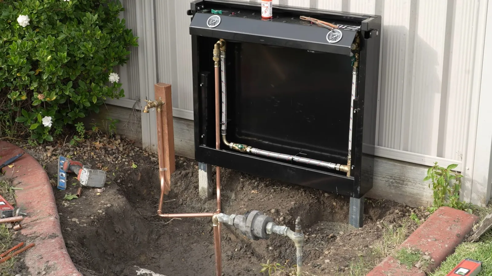 A black filter cabinet on legs against a metal-sided wall, with copper pipes and a meter in a dug-out bed below